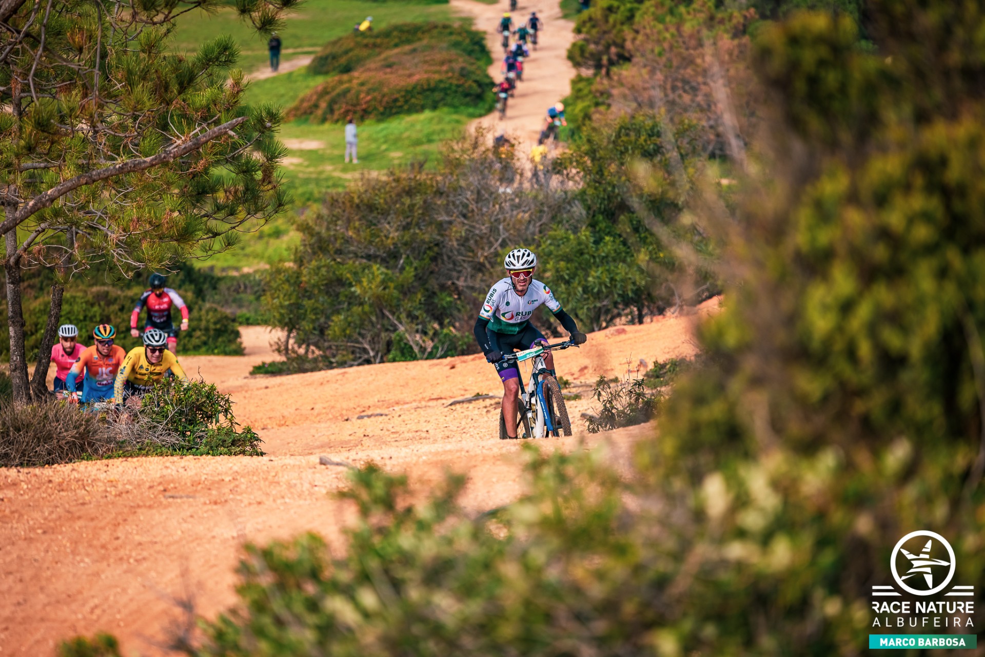 Ciclista com equipamento da Rubis Gás a percorrer um trilho de terra batida no RaceNature Albufeira, seguido por outros participantes.