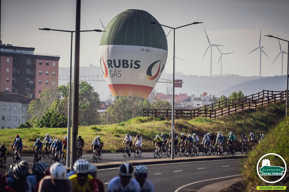Ciclistas a pedalar no Granfondo Torres Vedras, com um balão de ar quente da Rubis Gás a flutuar ao fundo.
