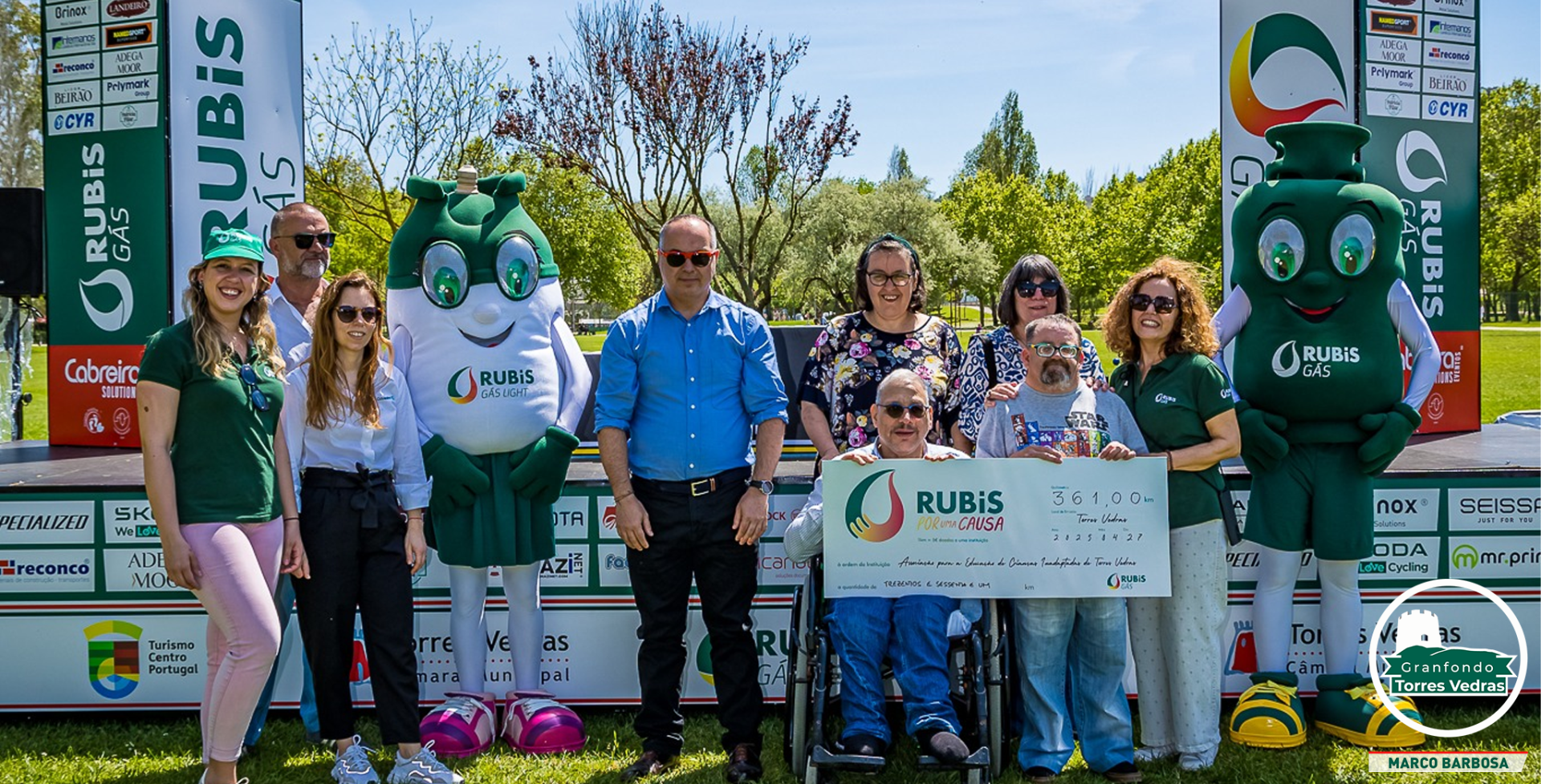 Entrega do cheque simbólico da iniciativa Rubis por uma Causa à APECI, com representantes da instituição e as mascotes da Rubis Gás num espaço ao ar livre.