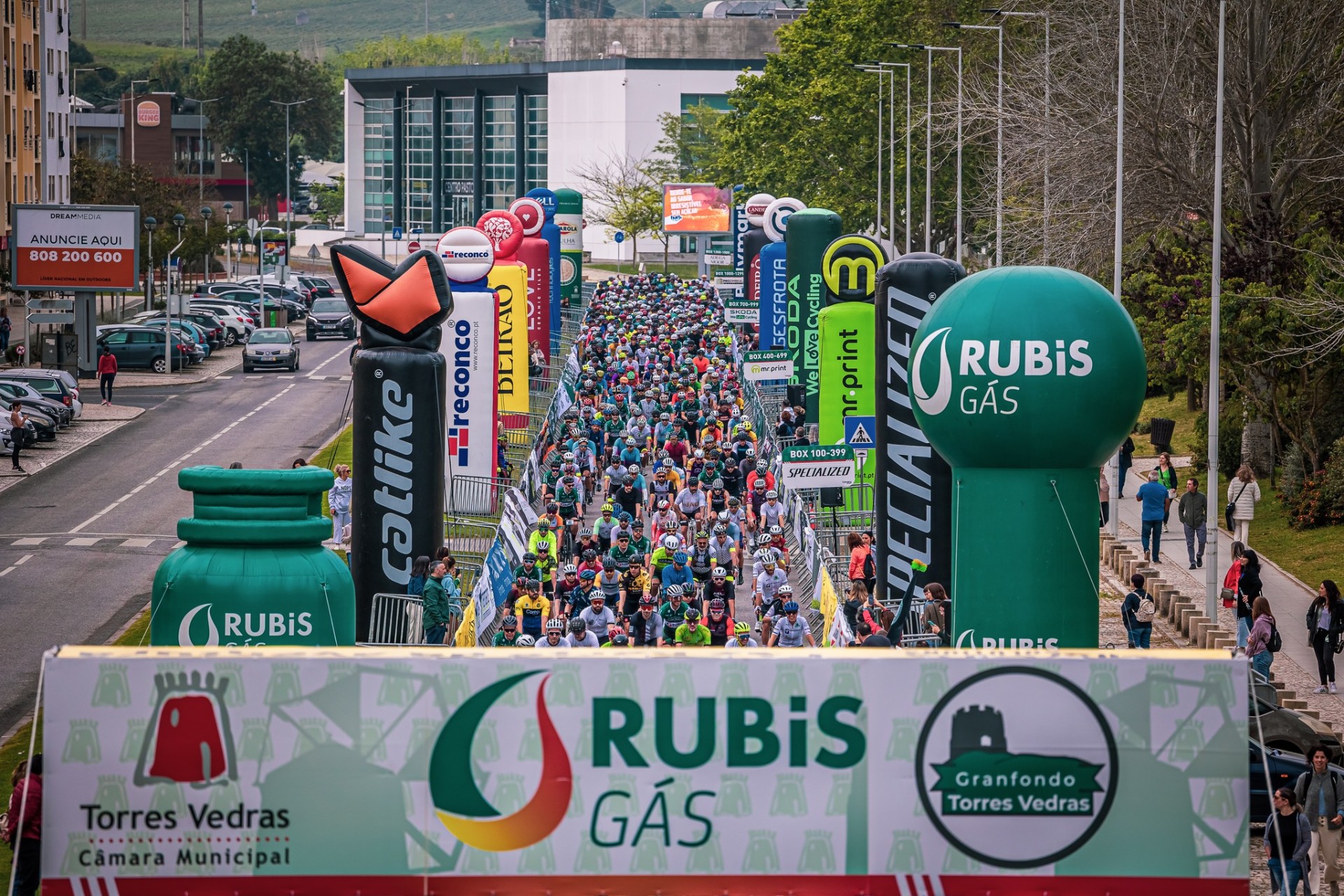 Grande pelotão de ciclistas alinhado no corredor de partida, ladeado por insufláveis publicitários da Rubis Gás.