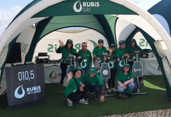 Grupo de utentes e representantes da CRACEP com t-shirts e bonés da Rubis Gás junto a bicicletas estáticas dentro da tenda da iniciativa.