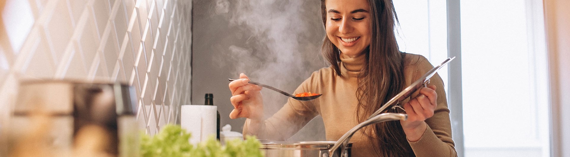Mulher a sorrir na cozinha enquanto levanta a tampa de uma panela a deitar vapor para provar a comida.