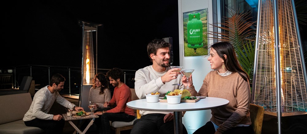 Pessoas a conviver numa esplanada à noite aquecida por aquecedores a gás, com um casal em destaque a brindar com cocktails