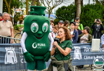 Representante da Rubis Gás a falar ao microfone ao lado da mascote verde da marca perante o público