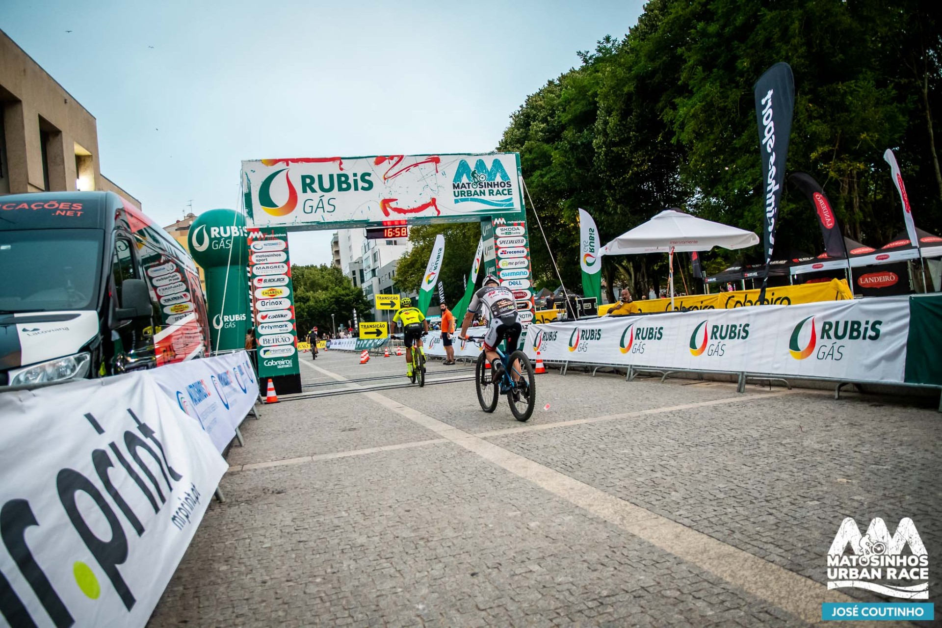 Vista do percurso urbano com ciclistas em ação a atravessar a meta do Matosinhos Urban Race, ladeada por lonas e insufláveis da Rubis Gás.