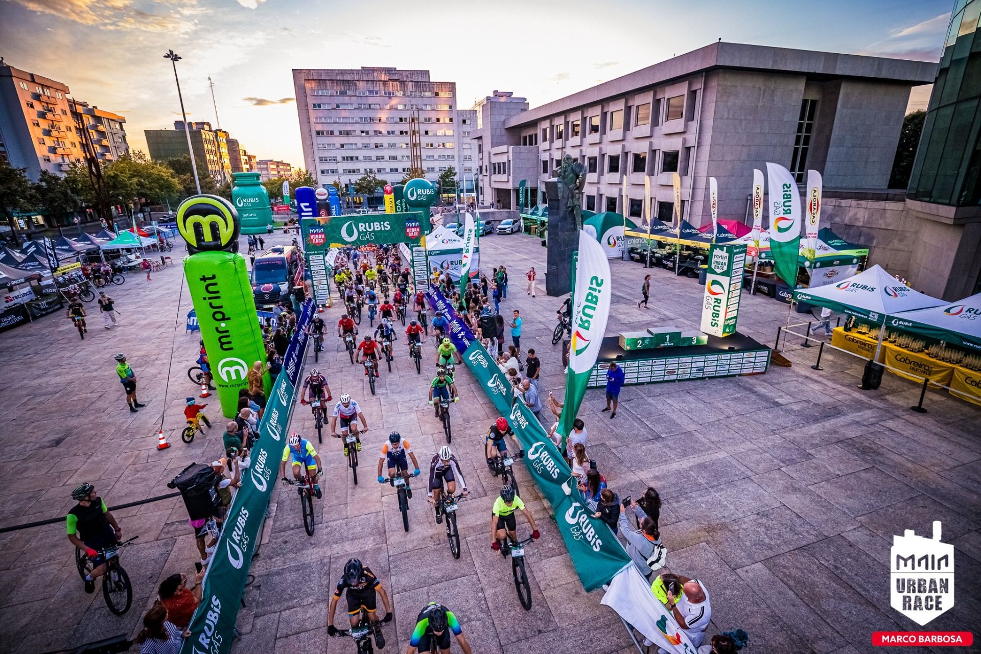 Vista superior do arranque dos ciclistas no Maia Urban Race, a percorrer o corredor delimitado por insufláveis e vedações da Rubis Gás.